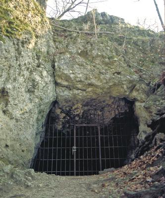 Aufwärts gerichteter Blick auf den vergitterten Eingang zu einer Höhle. Die Höhle befindet sich zwischen massigen, links etwas vorstehenden grauen Felsbänken.