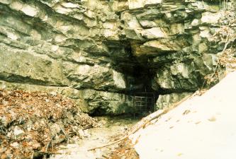 In massigen, grünlich grauen Felsbänken öffnet sich rechts der Bildmitte eine enge Höhle. Der Eingang ist mit einem Gitter gesichert. Aus der Höhle fließt Wasser in einem schmalen Bett auf den Betrachter zu.