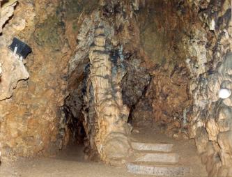 Blick in eine von Lampen erhellte Tropfsteinhöhle. Vor einer Nische, neben drei Treppenstufen, ragt ein leicht nach rechts gebogener großer Stalagmit vom Boden bis zur Decke.