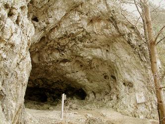 Das Bild zeigt eine fahl und gebleicht wirkende Felswand. Links öffnet sich der gewölbte Eingang einer Höhle. Ein gangbarer Weg mit Hinweistafeln führt hinein.