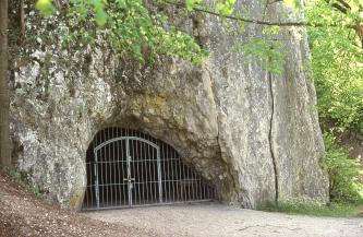 Das Bild zeigt den mit einer Gittertür verschlossenen halbrunden Eingang zu einer Höhle am Fuß einer steil aufragenden Felswand. Zum Eingang führt ein geebneter Weg.