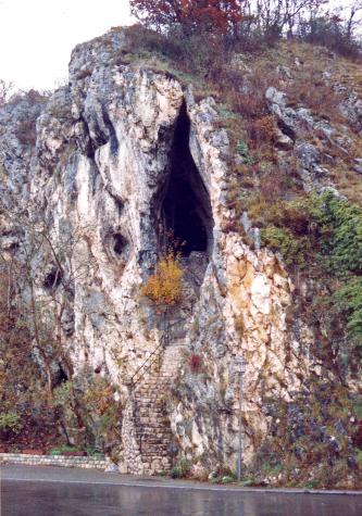 Blick auf eine steil aufragende Felsformation, deren rechte Flanke von Moos, Sträuchern und Bäumen bewachsen ist. Im hellen Mittelteil klafft die spitz zulaufende Öffnung einer Höhle. Eine Steintreppe führt aufwärts zum Eingang. Vorne fließt ein Fluss.