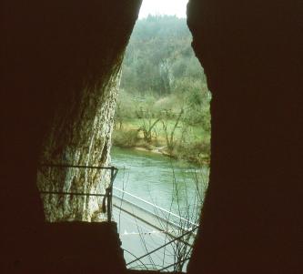 Blick aus einem schmalen, hohen Höhleneingang ins Freie. Die sichtbare Landschaft besteht aus einem Fluß und dessen Uferbereich sowie angrenzendem Wald.