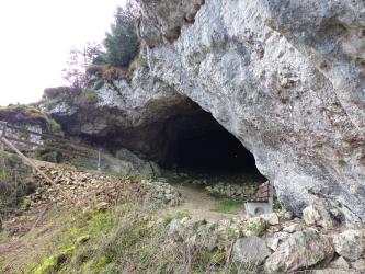 Seitenansicht einer nach links vorspringenden, weißlich grauen Felswand mit darunter liegender großer Höhlenöffnung. Entlang des Höhleneingangs verläuft ein gangbarer Weg. Vor und in der Höhle liegen Steinansammlungen.