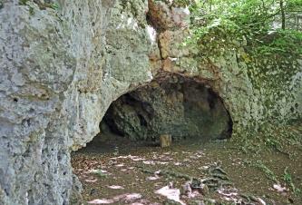 Blick auf die halbrunde Öffnung einer Höhle. Die den Eingang umstehenden Felswände sind links weißlich, rechts grünlich und dort auch bewachsen. Die Höhle lässt genug Licht herein, um auch hier Felswände erkennen zu können.