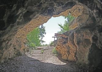 Blick aus der Vorhalle einer Höhle nach draußen. Wo Tageslicht auf den schräg liegenden, ovalen Eingang fällt, hat das Gestein eine rostbraune Farbe.