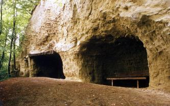 Leicht seitlicher Blick auf eine bräunlich graue Felswand, in der sich am Fuß zwei nischenartige Höhlen befinden. Die Höhle links ist fast rechtwinklig, die Höhle rechts ist höher und am oberen Ende gerundet. Eine Sitzbank ist darin aufgestellt.