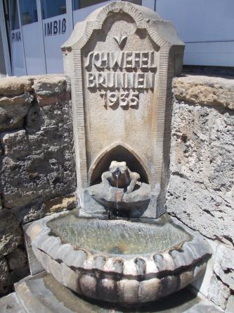 Im Eck einer Steinmauer steht ein ebenfalls aus Stein gefertigter Brunnen. Das Wasser, das aus einer froschartigen Figur fließt, gelangt in ein halbrundes, teilweise überlaufendes Becken.