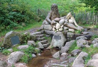 Blick auf eine kleine schmale Wasserquelle, von Steinblöcken gesäumt. An zweien dieser Blöcke sind Hinweistafeln angebracht. Der hintere Quellteil ist von einer großen Steinfigur verdeckt.