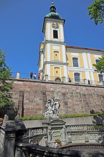 Im Vordergrund dieses Bildes ist die runde, steinerne Einfassung der Donauquelle zu sehen. Dahinter ragen eine hohe Mauer sowie eine mehrfarbige Kirche mit Glockenturm auf.