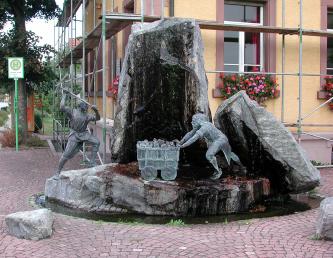 Blick auf einen steinernen runden Brunnen mit Figuren wie Bergarbeiter sowie einem kleinen beladenen Wagen. Zwei hohe und dicke Steinquader, einer stehend, einer halb liegend, stellen die Wände eines Bergwerks dar.