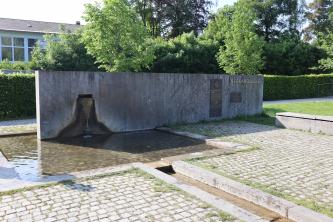 Blick auf die neu gestaltete Neckarquelle in Schwenningen mit Betonmauer, flachem Auffangbecken und schmalen Abflussrinnen. Der Quellausfluss erfolgt im oberen linken Teil der Mauer. Rechts sind mehrere Hinweistafeln an der Mauer angebracht.