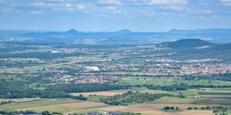 Blick von der Teck über das Albvorland nach Nordosten Weiter Blick von erhöhtem Standort auf eine flache bis hügelige Landschaft mit viel Wald und Wiesen, aber auch Äckern und Siedlungen. Im Mittelgrund sowie im Hintergrund erheben sich mehrere markante Berge.