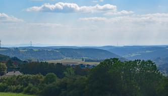 Blick aus größerer Höhe über Baumwipfel und Wald auf eine Stufenlandschaft mit flachen Rücken und Senken sowie bewaldeten Berghängen. 