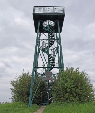 Blick auf einen Aussichtsturm mit graugrünem Metallgerüst, Wendeltreppe und offener Aussichtsplattform. Der Turm steht auf einer Anhöhe, sein Fuß ist von Laubbäumen umgeben.