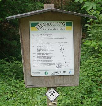 Blick auf eine kleine, überdachte Holztafel am Spiegelberger Bergbaulehrpfad. Die Tafel erzählt die Geschichte des Nassacher Kohlebergwerks und zeigt auch den Verlauf der verschiedenen Stollen.