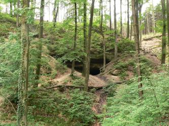 Blick auf einen im Wald gelegenen Felsenhang, an dessen unterem Ende sich der Eingang eines Stollens öffnet. Ein Wanderweg führt um den Hang herum.