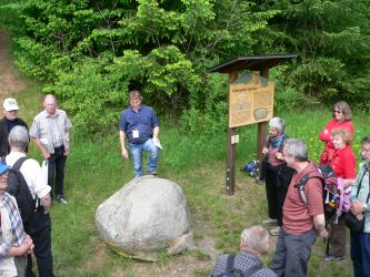 Weg der Kristalle, Waldbrunn Auf einer Wiese, links von einer Schautafel, liegt ein heller runder Felsblock. Ein Mann in blauem Hemd sagt gerade etwas über diesen Steinblock, und eine Besuchergruppe hört und sieht ihm dabei zu.