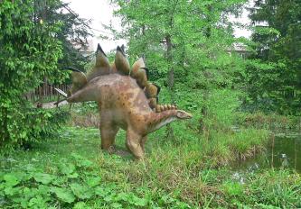 Das Bild zeigt die Nachbildung eines Sauriers im Außenbereich des Urwelt-Museums Hauff. Das gezeigte Tier geht auf vier Beinen und trägt dicke, abstehende Hornplatten auf dem Rücken. Neben Grünpflanzen und Bäumen gibt es rechts auch einen kleinen Teich.