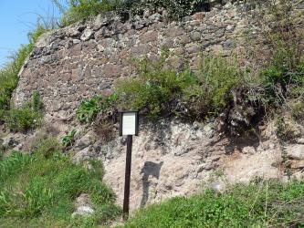 Blick auf gelblich graues Gestein, das am Fuß von Gras bewachsen ist. Über dem Gestein ist eine Stützmauer errichtet worden, aus grob zerkleinerten Steinbrocken. Dazwischen sowie oben wächst Gebüsch.