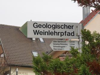 Blick auf einen Wegweiser mit grünem Pfeil, aufgestellt vor einer Häuserzeile. Das Schild zeigt zum Startpunkt des Geologischen Weinlehrpfads in Achkarren.