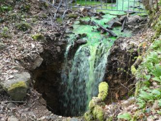Das Bild zeigt einen von rechts oben kommenden Bachlauf, dessen grün eingefärbtes Wasser in eine sich im Vordergrund öffnende Erdspalte fällt.