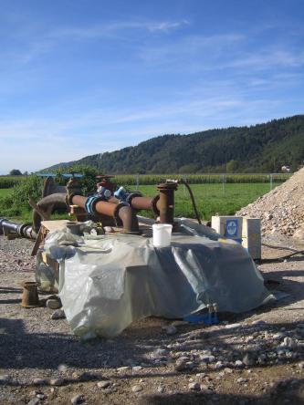 Das Bild zeigt mehrere Rohre, aufgebaut im Freien für einen Pumpversuch. Die Rohre liegen auf einem Holzgerüst, das im vorderen Teil mit einer Plane bedeckt ist. Rechts ist ein Abraumhügel erkennbar.