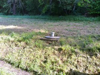 Das Bild zeigt eine viereckige Schachteinfassung mit Deckel, zum Sammeln einer Quelle. Der Schacht befindet sich auf ebenem, zum Vordergrund hin leicht abfallenden Gelände.
