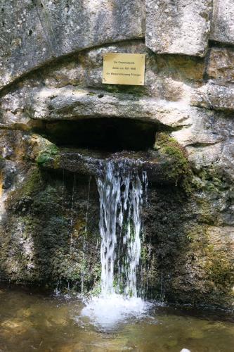 Aus einer quer liegenden Felsspalte fließt Wasser in einen Bach. Über der Spalte ist am Bildrand oben noch ein gemauerter Steinbogen sowie eine Schrifttafel erkennbar.
