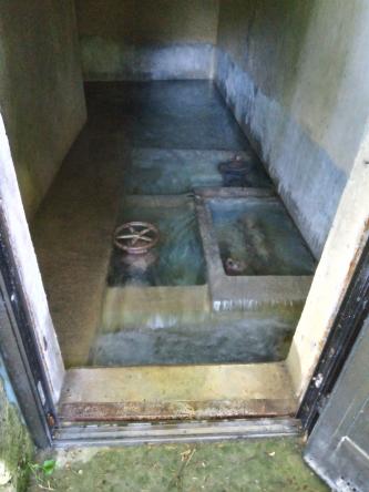 Leicht nach unten gerichteter Blick auf ein Wassersammelbecken mit hohen Einfassungswänden, Unterteilungen und Handrädern. Im Vordergrund ist ein türartiger Durchgang.