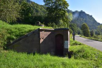 Blick auf ein kleines rotbraunes Gebäude mit Flachdach. Das Gebäude steht auf einem nach links aufsteigenden Wiesenhang mit Bäumen. Rechts führt eine Straße vorbei. Am Hang über dem Haus ist ein Schacht mit Deckel erkennbar.