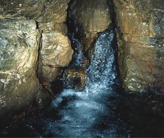 Zwischen eng stehendem, dunklem Felsgestein tritt Wasser hervor.