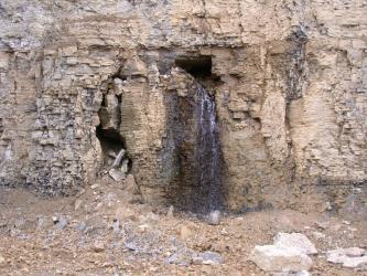 Das Bild zeigt eine hellbraune und zum Teil gräuliche Steinwand, in deren Mitte Wasser aus einem Hohlraum fließt.