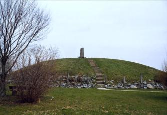 Auf dem Bild ist ein mit Gras bewachsener Grabhügel zu sehen. Auf den Hügel hinauf führt eine Steintreppe, oben steht ein Monument. Im linken Vordergrund befindet sich ein kahler Baum.