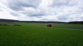 Die Rauhe Wiese bei Böhmenkirch-Heidhöfe – Blick nach Westen über die flache, mit Feuersteinlehm plombierte Karstwanne Blick über eine flachhügelige Wiesenlandschaft, die im Hintergrund von Ackerstreifen unterbrochen sowie von bewaldeten Bergrücken begrenzt wird. Entlang der Bergrücken sind mehrere Windräder zu erkennen.