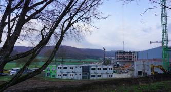 Bautätigkeit in der Siedlungsgasse des Kocher-/Brenz-Talzugs südlich von Oberkochen Blick über eine flachhügelige Wiesenlandschaft mit angrenzenden bewaldeten Bergen. Im Vordergrund werden mehrere Neubauten errichtet; zu sehen sind Baucontainer und Kräne. Links ragt ein verästelter Baum ins Bild.