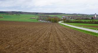 Riesalb bei Neresheim-Kösingen – Im Vordergrund mit erodierten Parabraunerden aus lösslehmreichen Fließerden über Bunter Brekzie (o64) Blick über eine höher gelegene, leicht abschüssige Ackerfläche. Im Mittel- und Hintergrund folgt eine flachhügelige, ansteigende Landschaft mit Wiesen, Äckern und Waldstreifen. Rechts neben einer kurvigen Straße liegt eine Ortschaft.