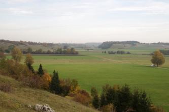 Blick einen Hang abwärts auf eine sich nach rechts ausbreitende flache Acker- und Wiesenlandschaft, die am seitlichen und hinteren Rand in flache bewaldete Hügel übergeht.