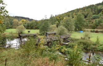 Das Bild zeigt eine hölzerne Brücke mit zwei seitlichen, überdachten Anbauten. Die Brücke führt quer über einen schmalen Fluss, dessen Ufer von Bäumen und Sträuchern eingefasst sind. Im Hintergrund folgen eine Wiese sowie bewaldete Berge.