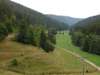 Blick in ein von einer Straße durchzogenes Tal, das links und rechts von bewaldeten Bergen gesäumt wird. Rechts sind an den Waldgrenzen flache grüne Wiesen zu sehen, links steigt das Gelände von der Straße her an und ist bräunlich, darüber auch rötlich.