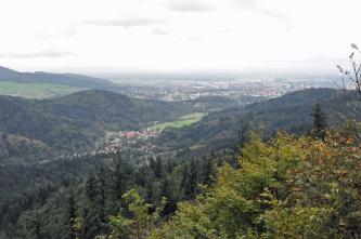 Blick aus großer Höhe über langegezogene bewaldete Bergrücken im Vorder- und Mittelgrund sowie eine weite, flache Ebene im Hintergrund. Die Ebene ist dicht besiedelt.