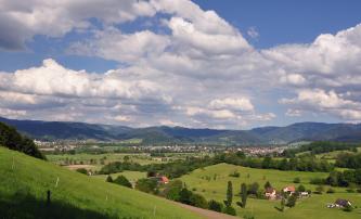 Vor einer bewaldeten Bergkette im Hintergrund breitet sich eine flache, rechts auch hügelige Ebene aus, die zu Füßen der Berge dicht, im Vordergrund dünner besiedelt ist. In der vorderen Mitte quert ein Waldstreifen die Ebene.