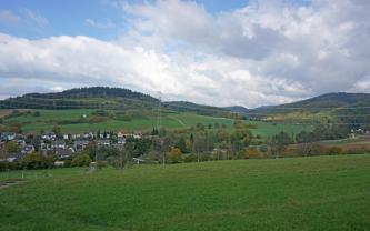 Zwischen grünen Hügeln im Vordergrund und teils grünen, teils bewaldeten Hügeln und Bergen im Hintergrund liegt ein schmales, links besiedeltes Tal.