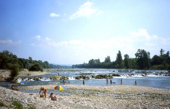 Das Bild zeigt einen breiten, von zahlreichen kleinen und großen Felsen unterbrochenen Fluss. Den Vordergrund begrenzt ein steiniger Uferbreich. Mehrere Menschen baden in dem Fluss.