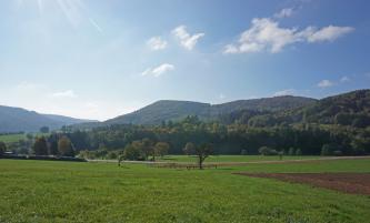 Im Vordergrund dieses Bildes sind flache Wiesen zu sehen, mit einer leichten Senke in der Bildmitte und einem Ackerstück rechts am Rand. Zum Hintergrund hin erheben sich bewaldete Hügel und Berge.