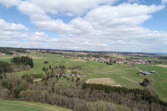 Blick von oben auf eine weit ausgebreitete, flache Landschaft mit Äckern, Wiesen, Wäldern und Siedlungen unter wolkigem Himmel. 