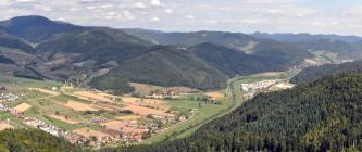 Von erhöhtem Standpunkt aus blickt man auf weite Nadelwälder rechts und eine aus landwirtschaftlichen Nutzflächen und Siedlungen bestehende Talebene links, durch die ein begradigter Fluss fließt. Dahinter folgt eine bewaldete Berglandschaft.