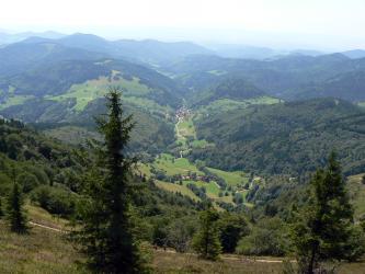 Aus großer Höhe blickt man in ein kleines, schmales Tal mit einer Siedlung am Ende. Links und rechts des Tales sowie im Vorder- und Hintergrund erheben sich steile, teilweise bewaldete Hügel und Berge.