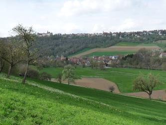 Der Blick geht über eine nach unten hin abfallende grüne Wiese über ein bewirtschaftetes Tal, an das sich ein wieder ansteigender, teils bewaldeter Hang anschließt. Im Mittelgrund sowie auf der Anhöhe liegen Ortschaften, hinten links auch ein Schloss..