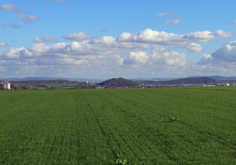 Blick über eine hochgelegene grüne Ackerfläche auf ein angrenzendes, weites flaches Tal. Hier sind neben Siedlungen auch ein grüner Bergkegel mit einem Windrad zu sehen. Im fernen Hintergrund verlaufen bewaldete Bergrücken.